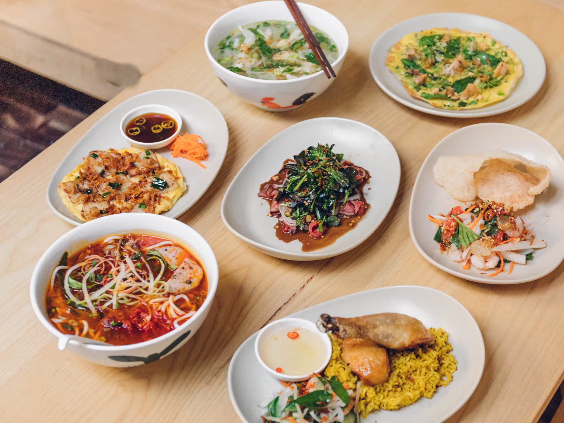 Seven dishes from Đi Ăn Đi arranged on a table. Starting from the bottom left corner and moving clockwise: Bún Bò Huế (Spicy Lemongrass Beef Noodle Soup), Banh Bot Chien (Fried Daikon Rice Cake Omelette), Phở Gà (Chicken Noodle Soup), Banh Trang Nuong So (Vietnamese “Pizza” With Clams), Gỏi Bưởi (Pomelo Salad With Rock Shrimp) and chicken with rice. In the center of the table is Bo Thai Chanh (Beef Carpaccio).